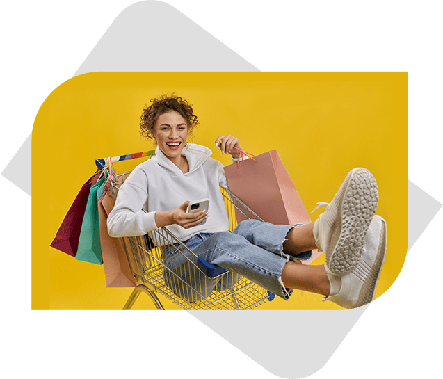 A smiling girl sits inside a shopping cart, holding a shopping bag in one hand and a mobile phone in the other, against a vibrant yellow background.